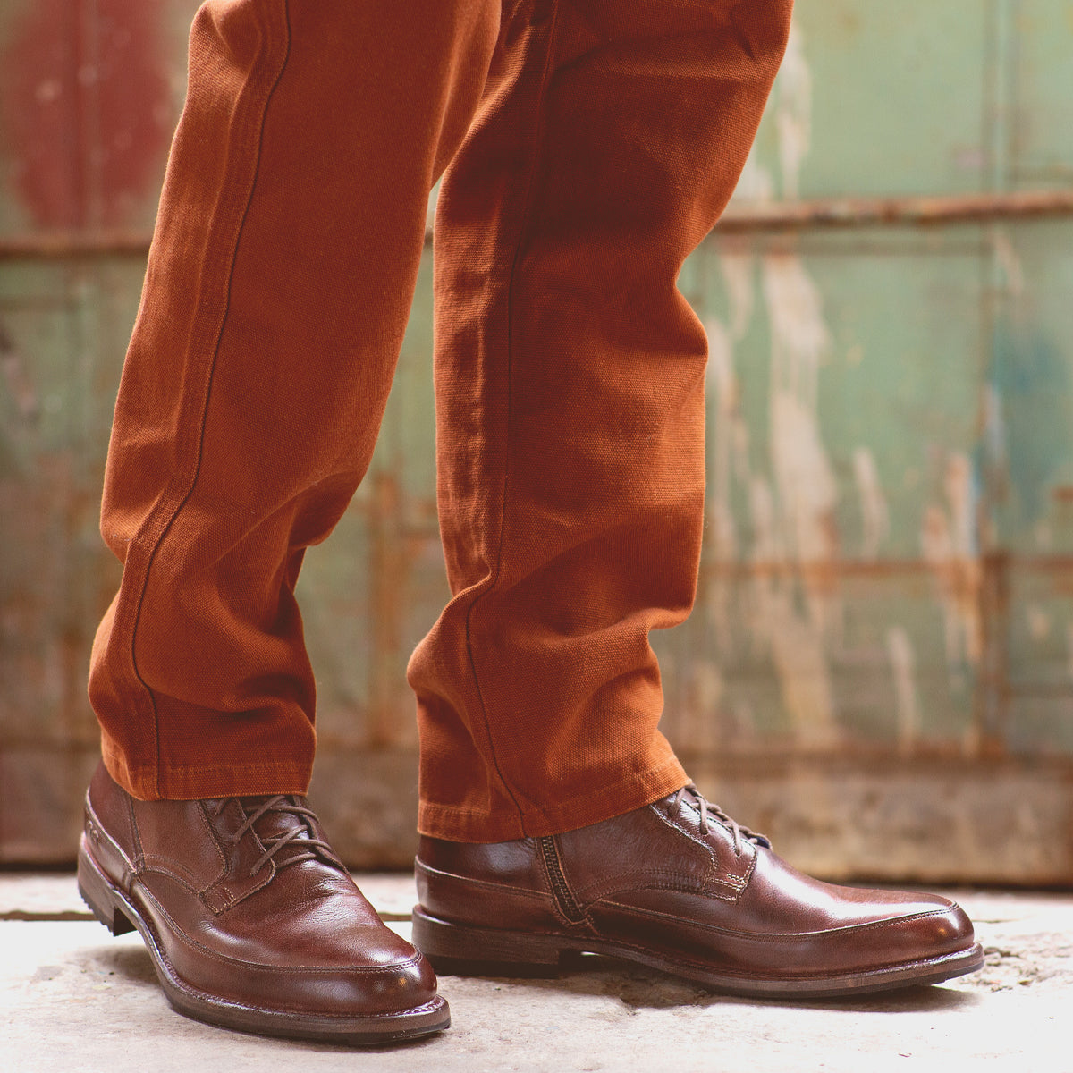 Close-up of a person's lower legs wearing brown corduroy pants and Bed Stu polished brown leather lace-up ankle boots, standing in front of a weathered metal backdrop.