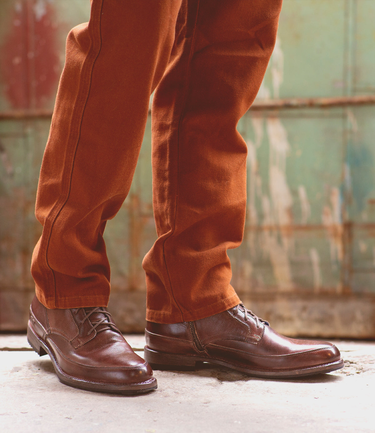 Close-up of a person's lower legs wearing brown corduroy pants and Bed Stu polished brown leather lace-up ankle boots, standing in front of a weathered metal backdrop.