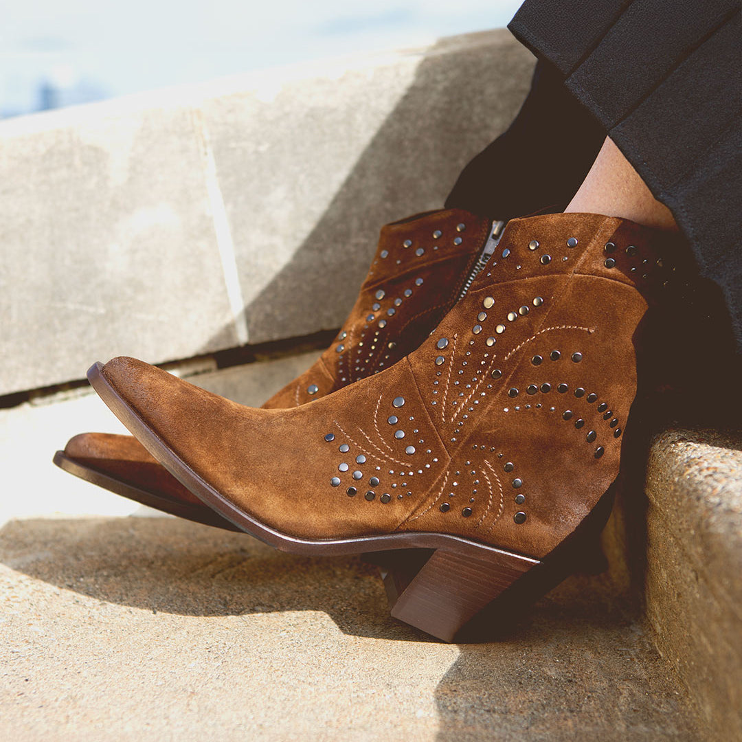 The person sitting on stone steps is wearing the Sposa by Bed Stu, featuring brown suede ankle boots with a leg-lengthening effect, decorative stitching, and studded details.