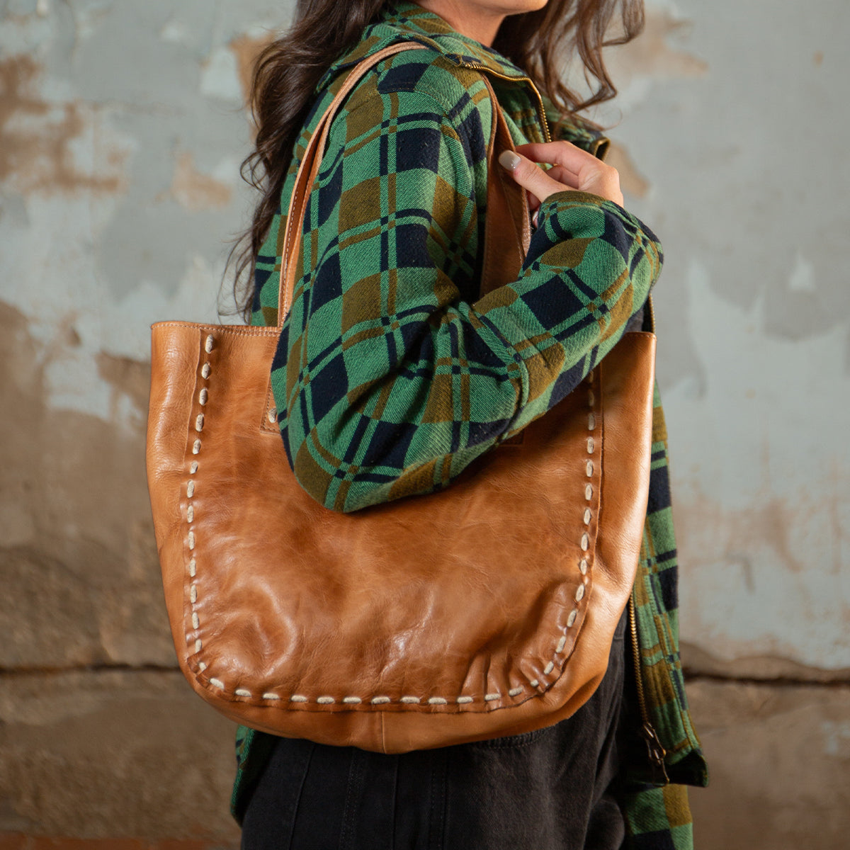 A woman in a green and black plaid shirt carries the Bed Stu Stevie tote, featuring leather handles and visible edge stitching for a vintage-chic modern look.
