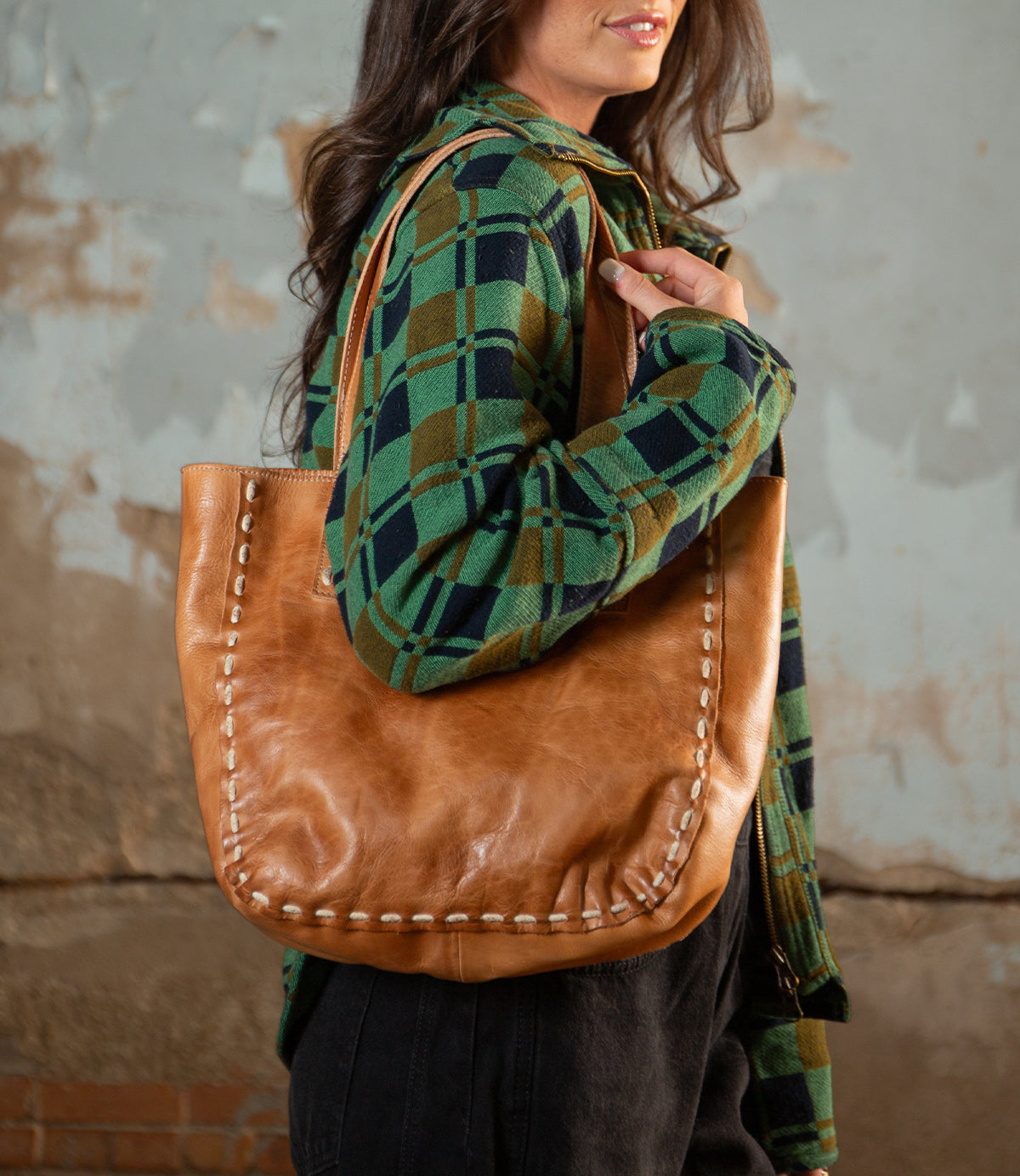 A woman in a green and black plaid shirt carries the Bed Stu Stevie tote, featuring leather handles and visible edge stitching for a vintage-chic modern look.