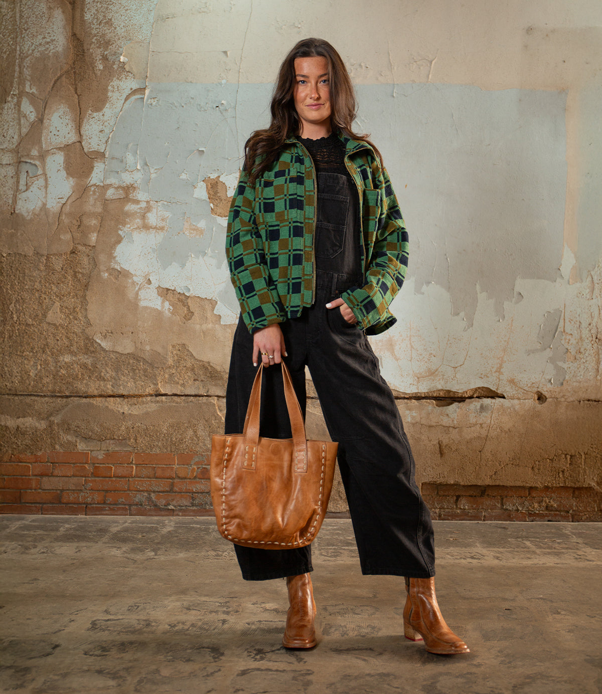 A woman stands indoors by a weathered wall, carrying the Stevie vintage-chic modern tote with leather handles from Bed Stu, wearing a green plaid jacket, black overalls, and brown boots.