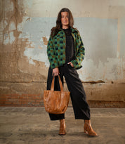 A woman stands indoors by a weathered wall, carrying the Stevie vintage-chic modern tote with leather handles from Bed Stu, wearing a green plaid jacket, black overalls, and brown boots.