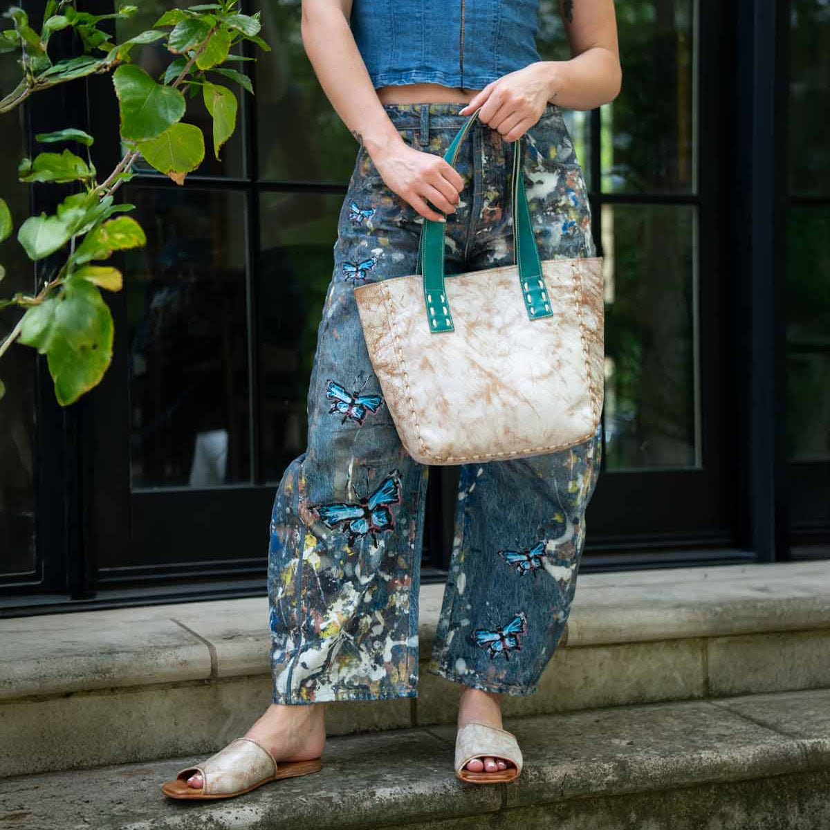 Outdoors, a person wears a sleeveless denim top, butterfly-painted jeans, and open-toe sandals, holding the Bed Stu Stevie—a vintage bag with green handles that's perfect for everyday carry.