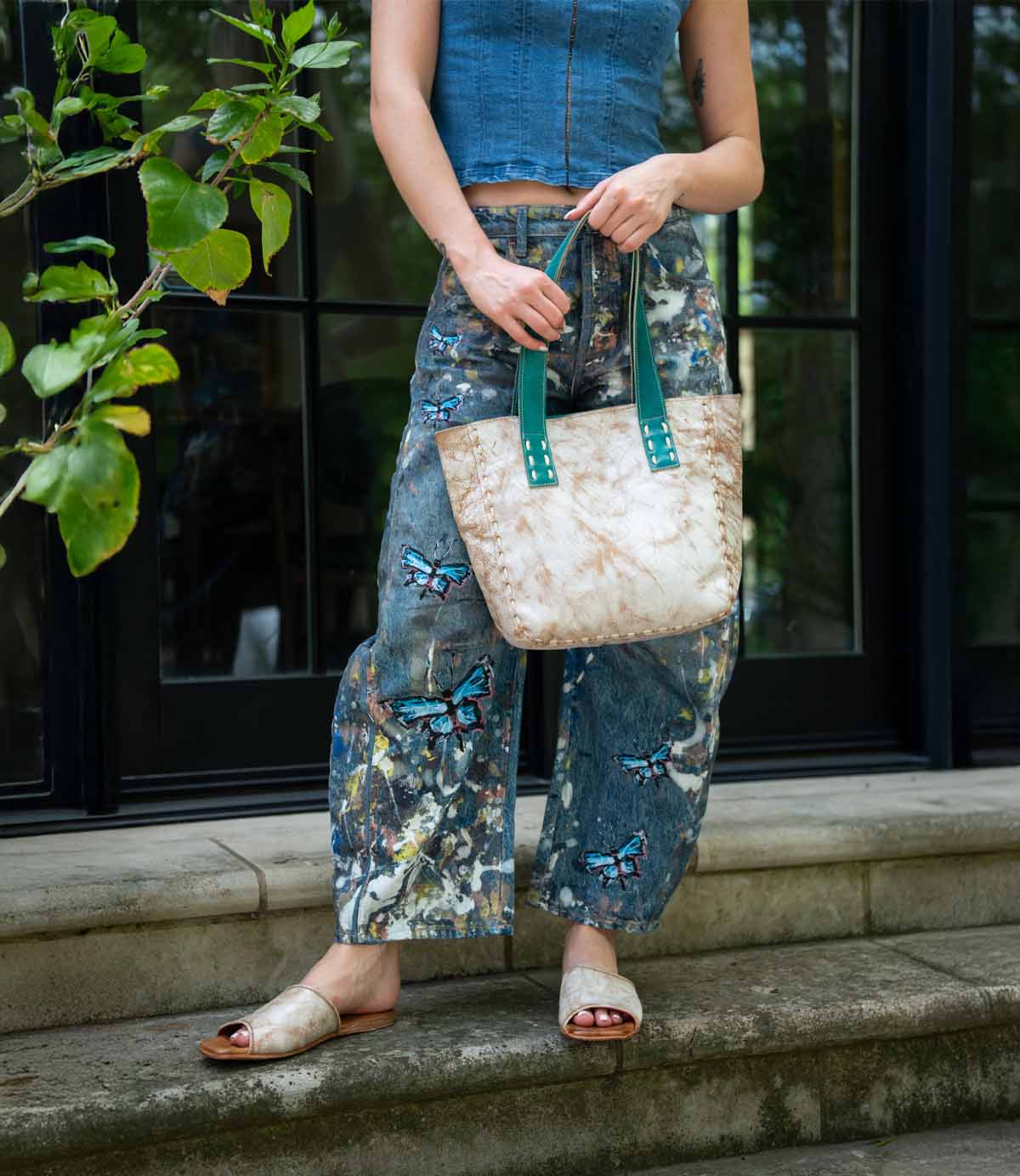 Outdoors, a person wears a sleeveless denim top, butterfly-painted jeans, and open-toe sandals, holding the Bed Stu Stevie—a vintage bag with green handles that's perfect for everyday carry.