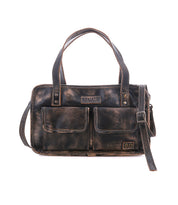The Bed|Stü Stuffy is a brown leather handbag featuring a spacious interior, two front flap pockets, dual handles, and an adjustable shoulder strap, shown against a white background.