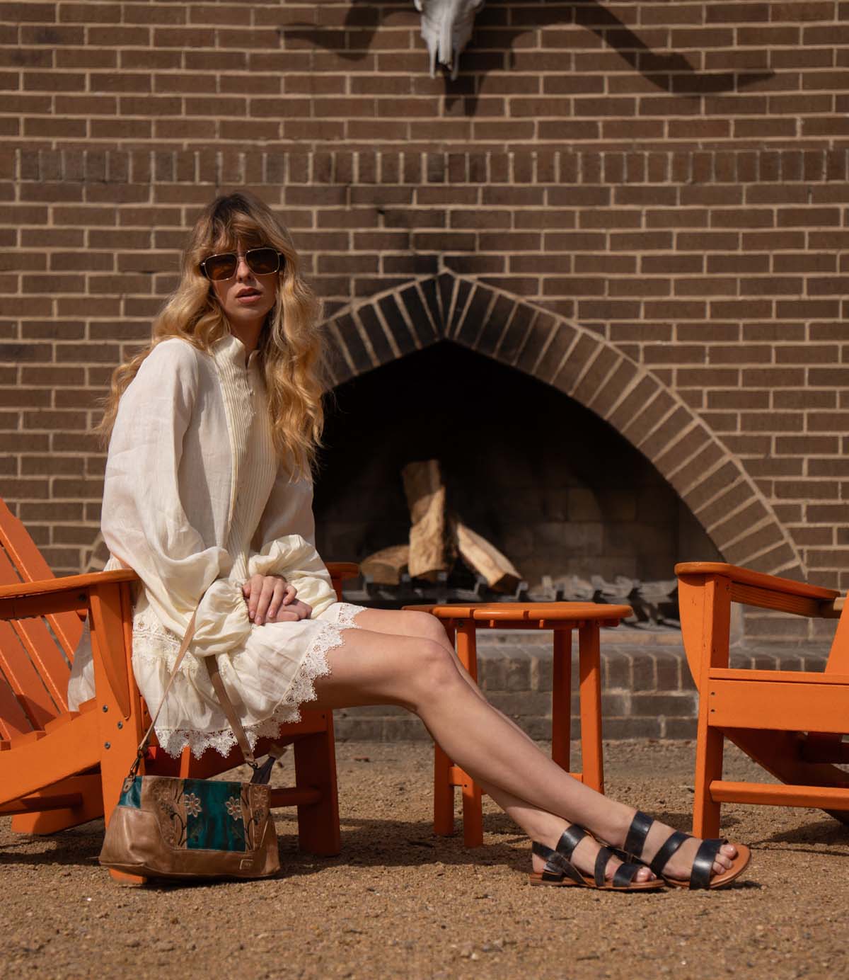 A woman wearing Bed Stu's Superb leather sandals with an adjustable ankle strap showcases effortless everyday style, seated in a white dress and sunglasses by a brick fireplace, her handbag beside her.