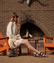 A woman wearing Bed Stu's Superb leather sandals with an adjustable ankle strap showcases effortless everyday style, seated in a white dress and sunglasses by a brick fireplace, her handbag beside her.
