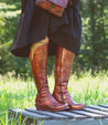 A woman in a black dress and brown boots with western-shaped heel standing on a bench, wearing the Takoma boots by Bed Stu.