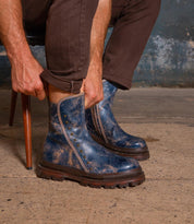 A person in brown pants sits on a chair, pulling on Bed Stu’s Tanto men's boots—blue, distressed leather with metal studs and a lug outsole—while on a concrete floor.