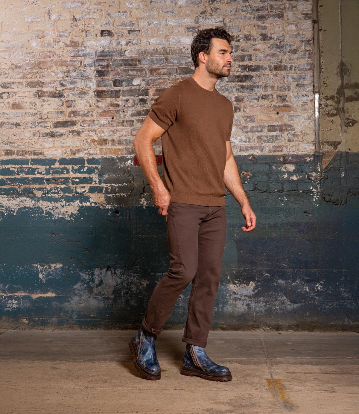 A man wearing brown pants, a brown short-sleeve sweater, and Bed Stu Tanto men's boots with rugged lug outsole stands before a worn painted brick wall, bringing European flair to the scene.