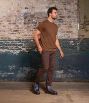 A man wearing brown pants, a brown short-sleeve sweater, and Bed Stu Tanto men's boots with rugged lug outsole stands before a worn painted brick wall, bringing European flair to the scene.
