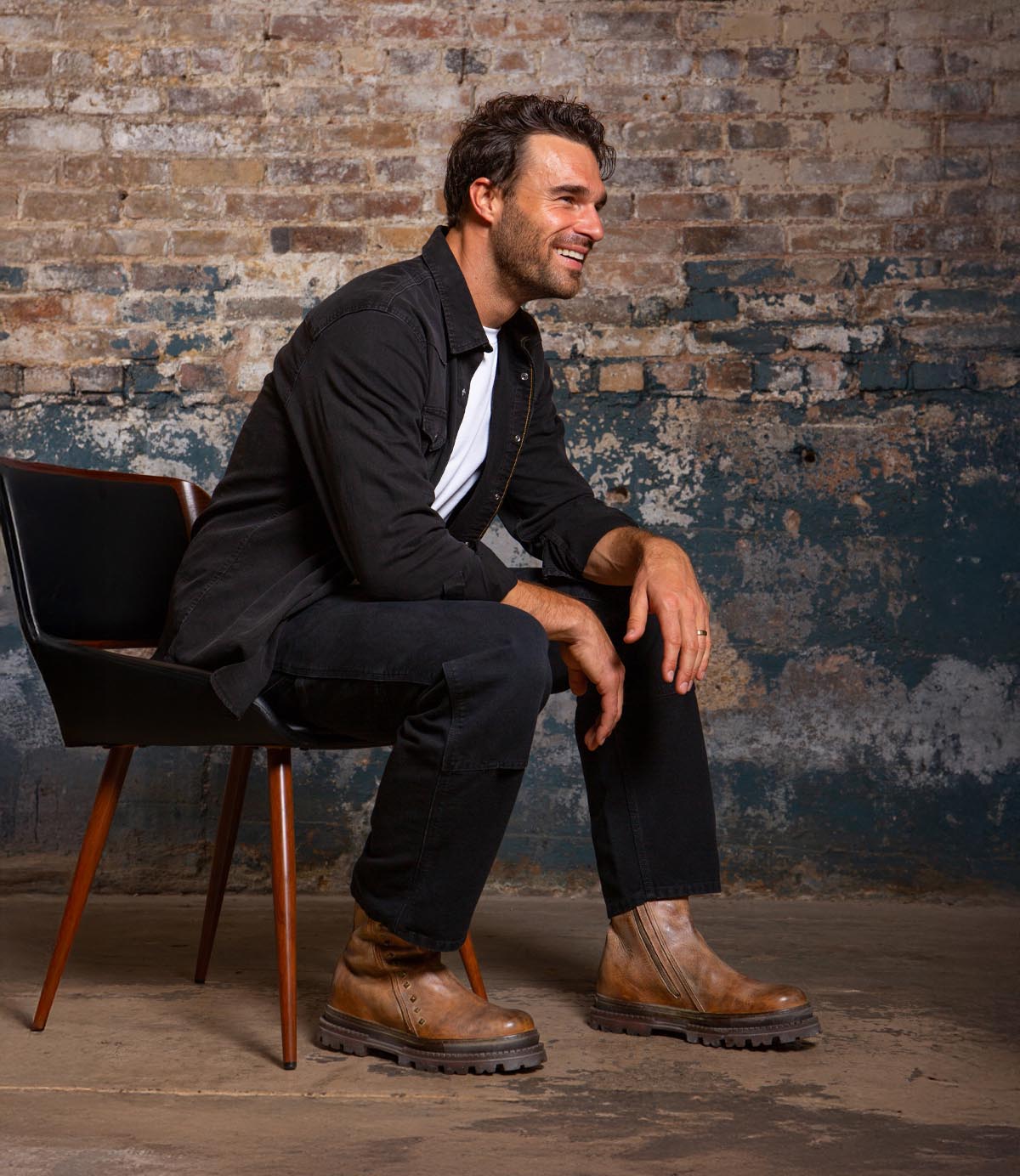 A man with short dark hair and a beard sits sideways on a black chair, dressed in black and wearing Bed Stu Tanto brown men's boots with lug outsoles, set against a distressed brick wall.