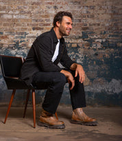 A man with short dark hair and a beard sits sideways on a black chair, dressed in black and wearing Bed Stu Tanto brown men's boots with lug outsoles, set against a distressed brick wall.