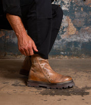 A person in black pants adjusts the Bed Stu Tanto men's boot, featuring metal studs and a rugged lug outsole, against a worn concrete floor and weathered wall for a subtle touch of European flair.