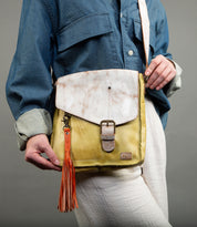 A person holds Bed Stu's "Tassel Clip," a yellow and white leather crossbody bag featuring a distinctive buckle and vibrant orange tassel. Paired effortlessly with a denim shirt and white pants, this versatile accessory complements any occasion.