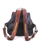 The Bed Stu Teji is a black statement backpack with brown leather straps, gold embroidery, a side zipper pocket, and silver hardware, photographed against a white background.