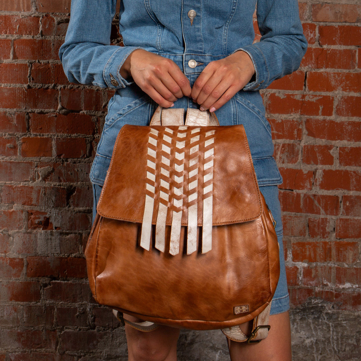 A person in a denim dress holds the Bed Stu Teji statement backpack with white geometric accents, standing before a red brick wall.