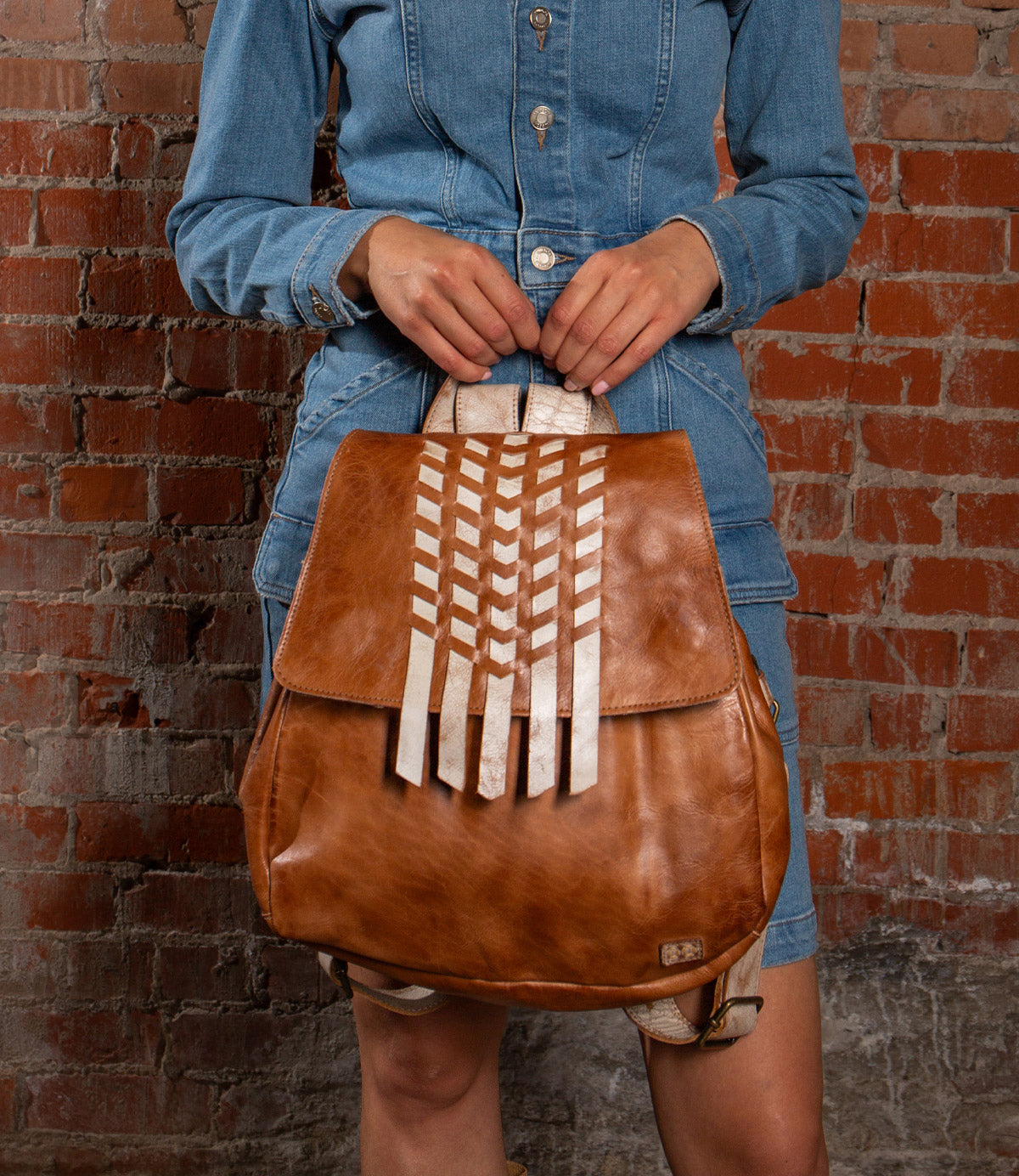 A person in a denim dress holds the Bed Stu Teji statement backpack with white geometric accents, standing before a red brick wall.