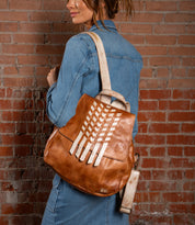 A woman in denim poses by a brick wall, carrying the Bed Stu Teji backpack—brown leather with beige decorative strips and a matching beige strap.