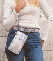 A person in a cream sweater and jeans holds the Templeton II, a blue marble-patterned crossbody handbag with a strap, by Bed Stu.