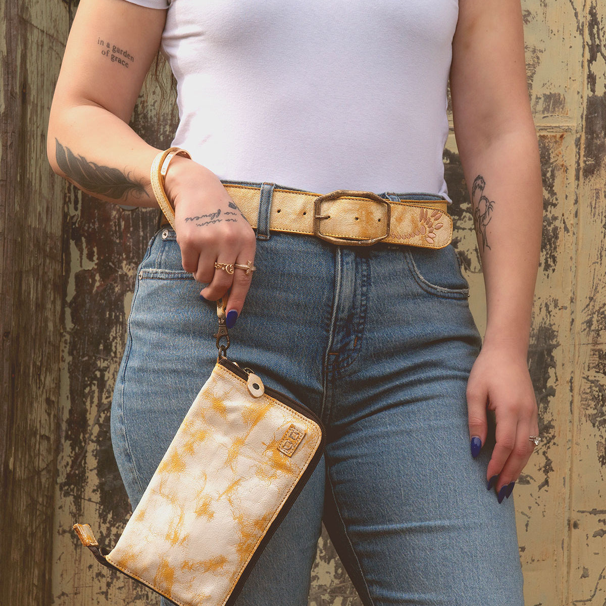 A person in a white t-shirt and jeans holds the Templeton II, a yellow patterned clutch by Bed Stu, perfectly paired with their matching belt and rings. Tattoos snake down their arm and hand against a rustic background.
