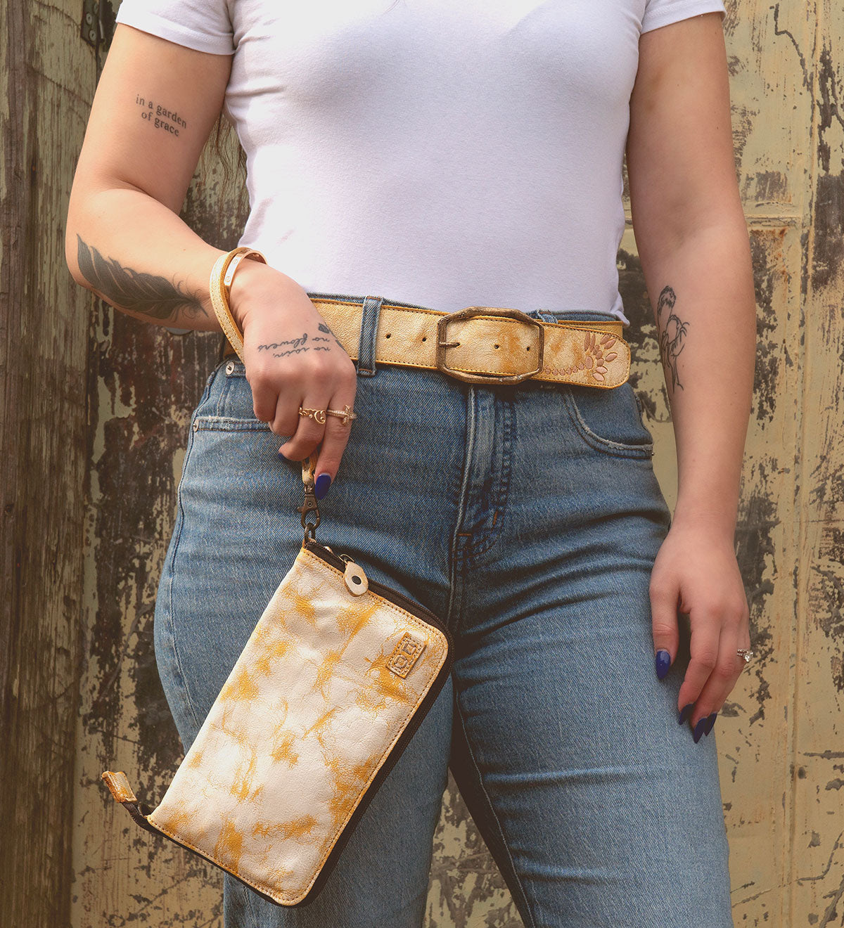 A person in a white t-shirt and jeans holds the Templeton II, a yellow patterned clutch by Bed Stu, perfectly paired with their matching belt and rings. Tattoos snake down their arm and hand against a rustic background.