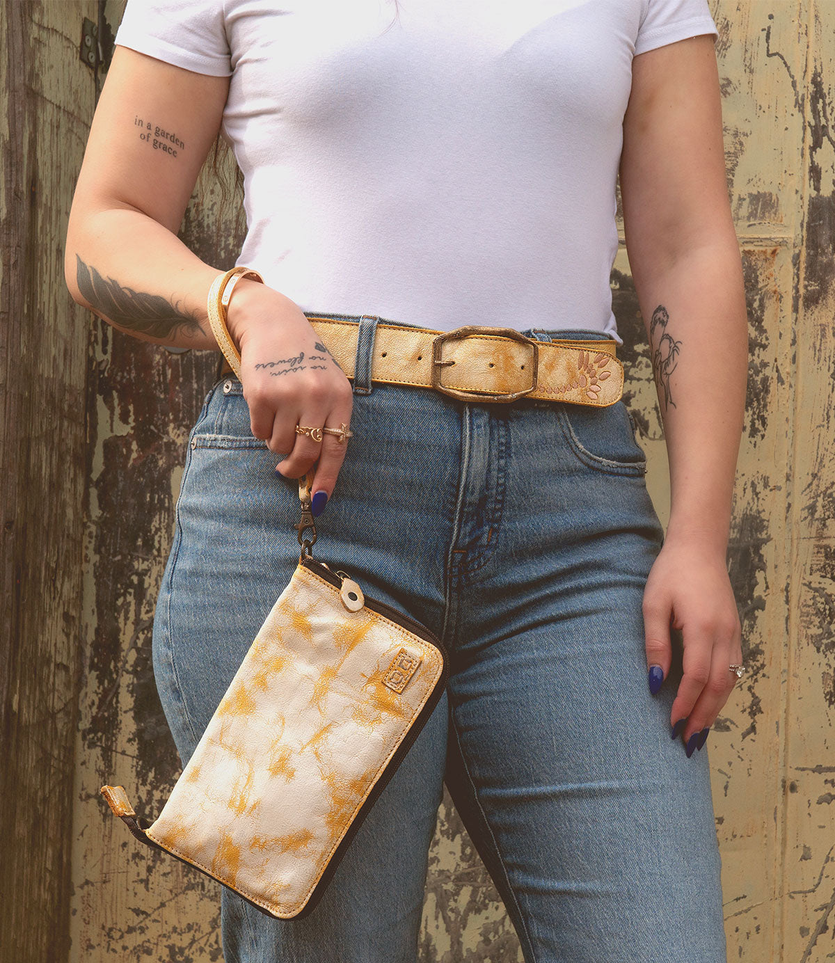 A person in a white t-shirt and jeans holds the Templeton II, a yellow patterned clutch by Bed Stu, perfectly paired with their matching belt and rings. Tattoos snake down their arm and hand against a rustic background.