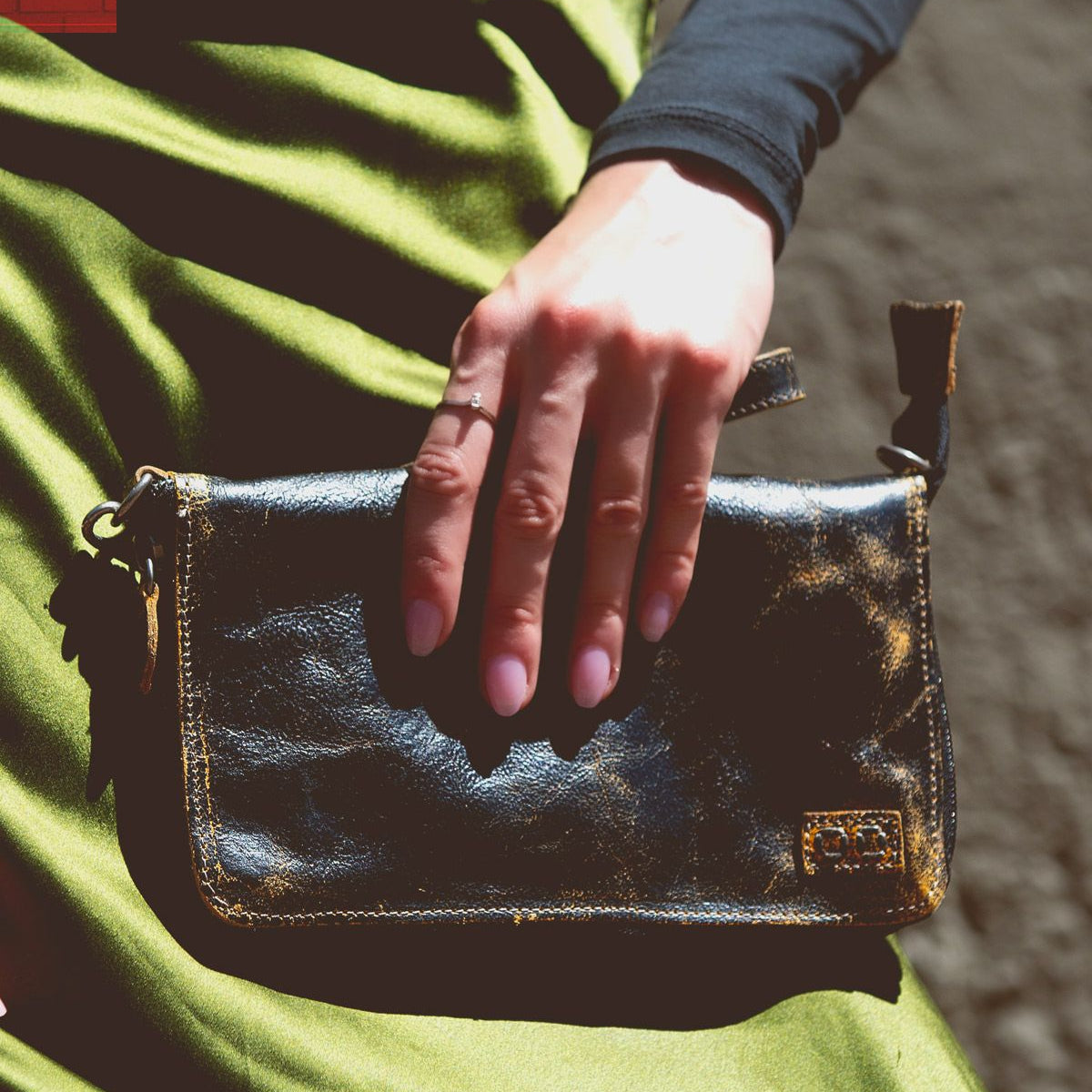 A hand rests on the Templeton II, a small black leather clutch by Bed Stu, against a green satin fabric, showcasing the elegance of this versatile accessory that could double as a chic crossbody.