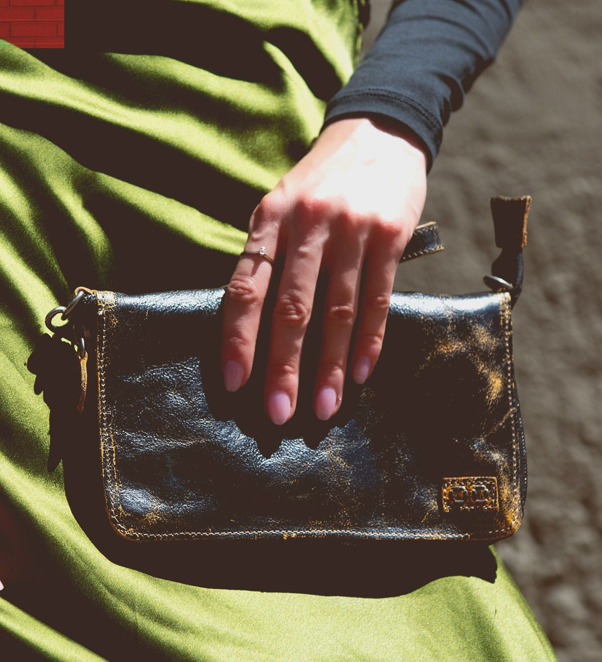 A hand rests on the Templeton II, a small black leather clutch by Bed Stu, against a green satin fabric, showcasing the elegance of this versatile accessory that could double as a chic crossbody.