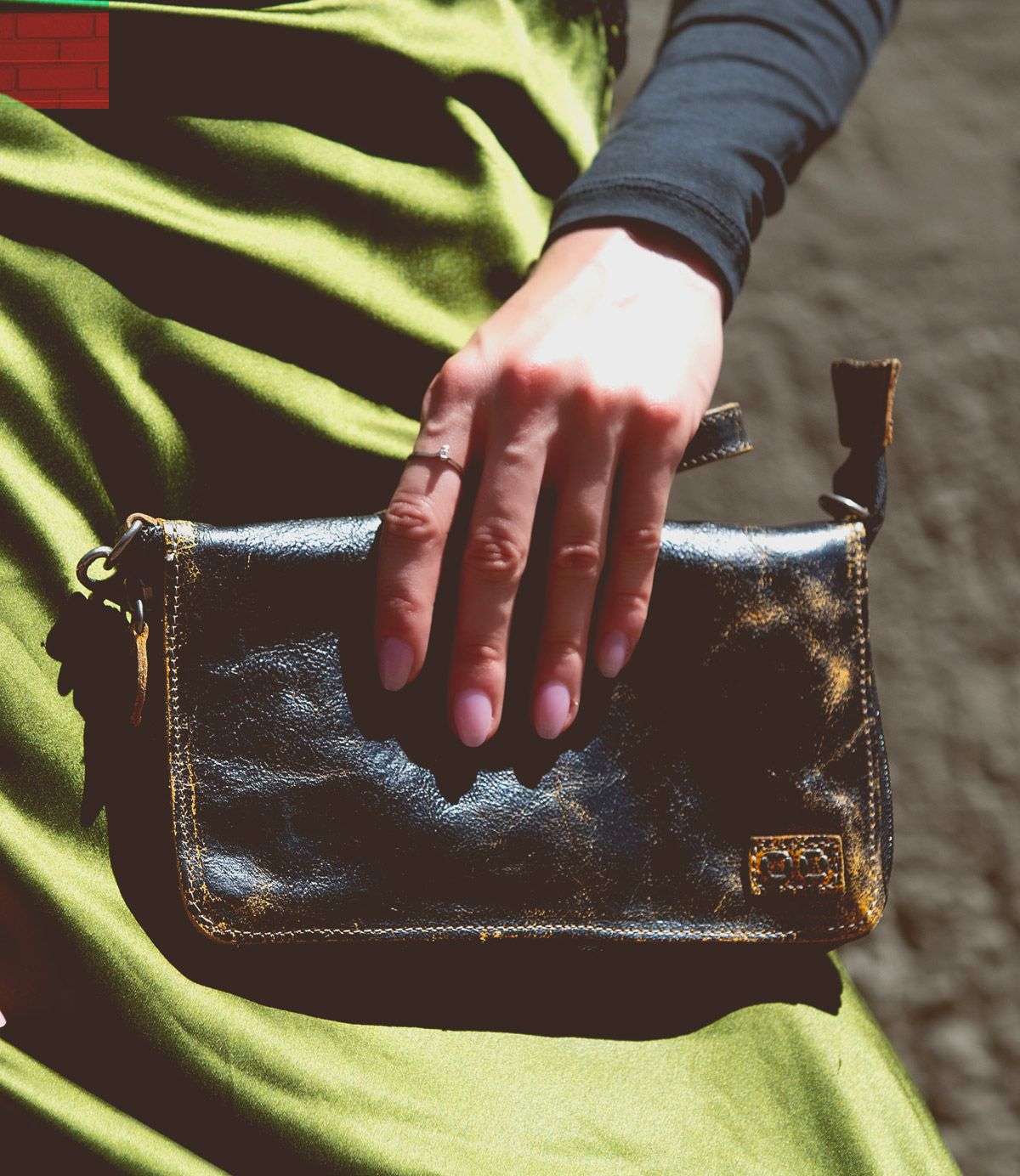 A hand rests on the Templeton II, a small black leather clutch by Bed Stu, against a green satin fabric, showcasing the elegance of this versatile accessory that could double as a chic crossbody.