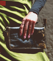 A hand rests on the Templeton II, a small black leather clutch by Bed Stu, against a green satin fabric, showcasing the elegance of this versatile accessory that could double as a chic crossbody.