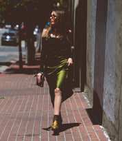 A woman wearing a green dress and black boots walks on a sunlit sidewalk while holding the Bed Stu Templeton II small leather wallet.