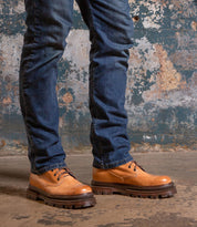 A person in blue jeans stands on a concrete floor by a weathered wall, wearing Bed Stu’s Tempt—a stylish hybrid shoe that blends the classic Chelsea boot look with rugged, thick soles.
