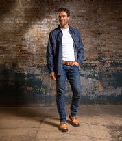 A man stands indoors against a worn brick wall, wearing a white t-shirt, blue denim shirt, jeans, a brown belt, and Bed Stu Tempt stylish hybrid Chelsea boots.