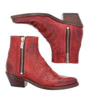 The Bed Stu Tentativa is a pair of red leather ankle boots featuring artisan detailing, decorative stitching, side zippers, and low stacked heels, shown from the side and bottom views.