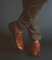 A person wearing handcrafted brown Time loafers by Bed Stu and olive green pants stands against a dark background, exuding sophistication.