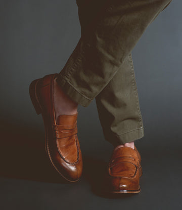 A person wearing handcrafted brown Time loafers by Bed Stu and olive green pants stands against a dark background, exuding sophistication.