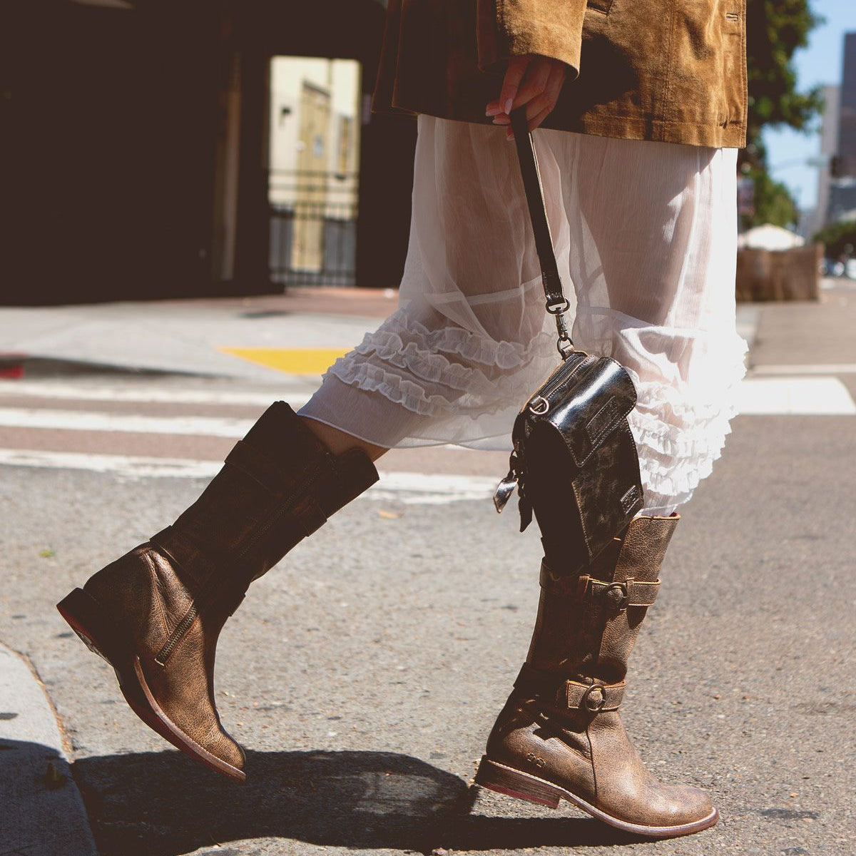 A person walking on the street is wearing a brown jacket, a sheer white dress with white ruffle trim, and Bed Stu's Turn brown leather boots made with Goodyear welted construction, while holding a small black handbag.
