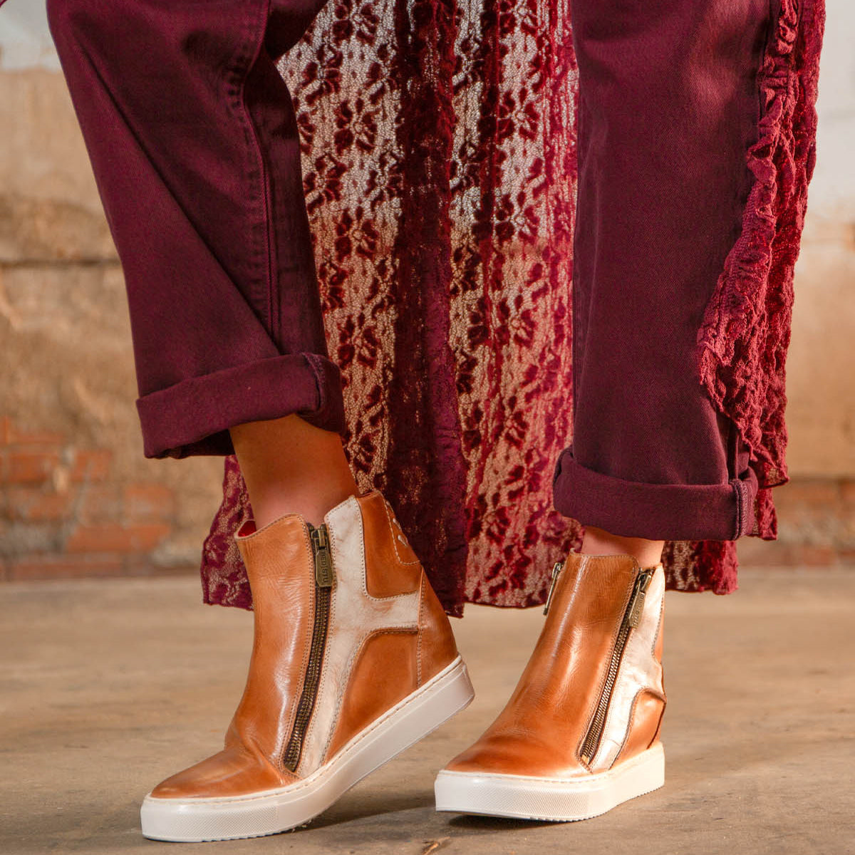 A person stands on a concrete floor wearing Bed Stu's Twist tan high-top sneakers with leather panels and side zippers, paired with burgundy pants and a textured burgundy outer layer.