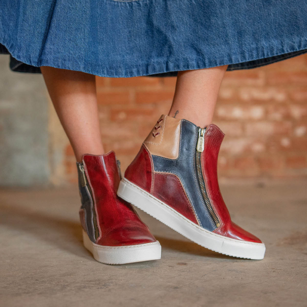 A person wears the Bed Stu Twist high-top sneakers featuring leather panels and side zippers, paired with a blue denim skirt, standing on a concrete floor before a brick wall.