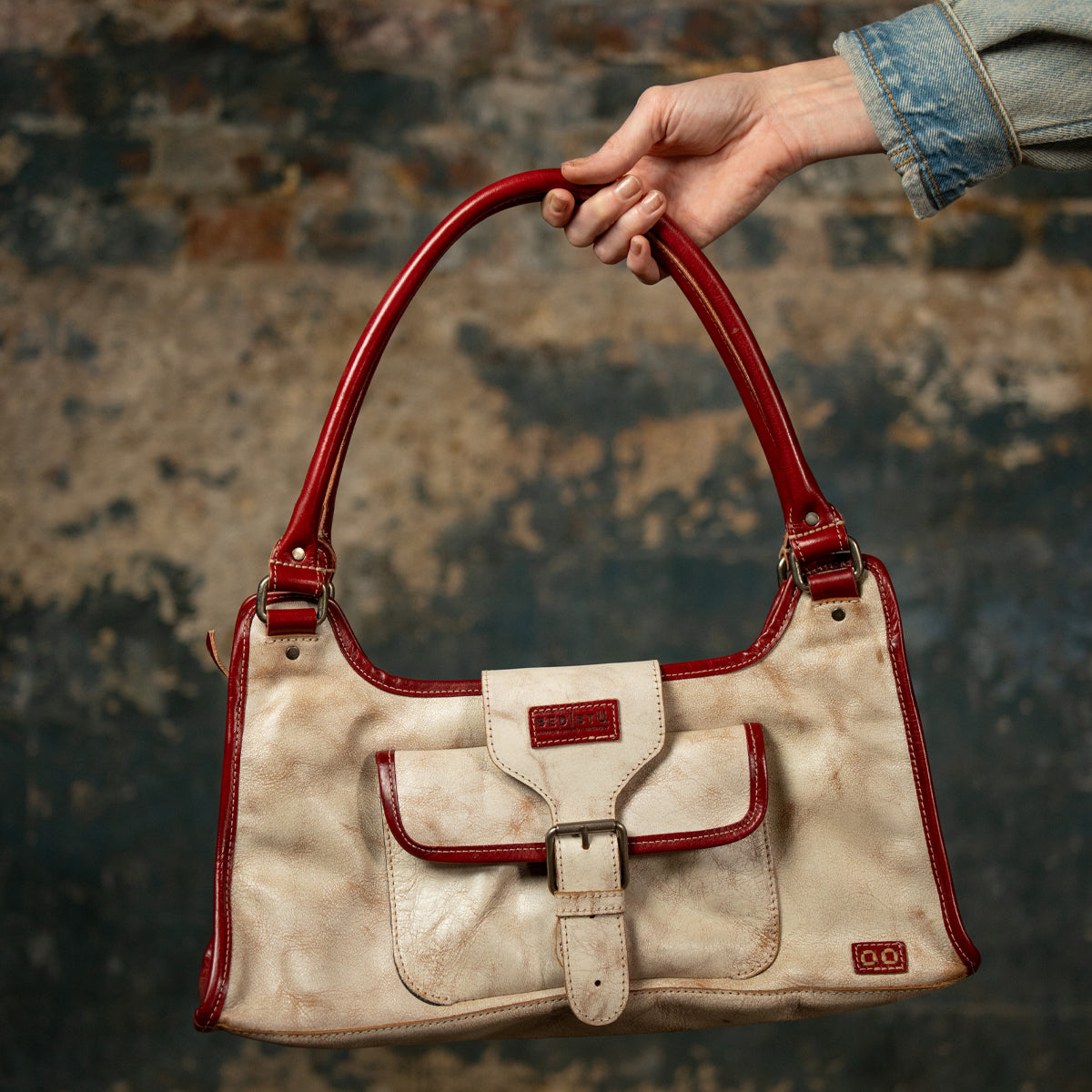 A hand holds the Bed Stu Unbending—a beige washed leather shoulder bag with red handles, trim, and a front pocket—against a textured stone background. An elegant work-bag for any occasion.
