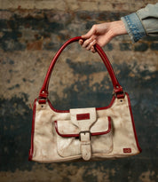 A hand holds the Bed Stu Unbending—a beige washed leather shoulder bag with red handles, trim, and a front pocket—against a textured stone background. An elegant work-bag for any occasion.