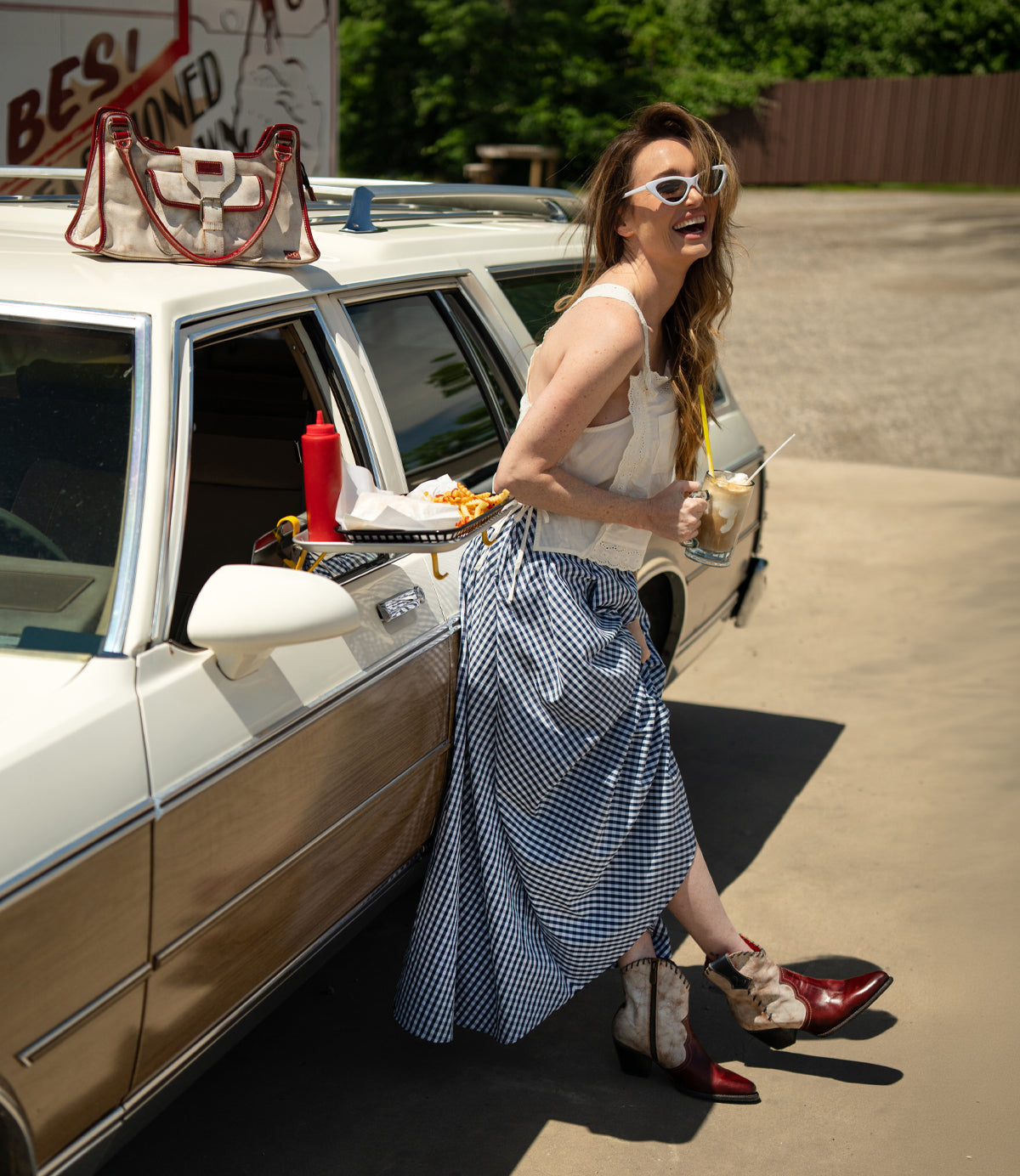 A woman in a gingham skirt and cowboy boots stands by a vintage car, holding a drink and fries, her Bed Stu Unbending washed leather shoulder bag at her side, with a tray of food attached to the car window.