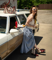 A woman in a gingham skirt and cowboy boots stands by a vintage car, holding a drink and fries, her Bed Stu Unbending washed leather shoulder bag at her side, with a tray of food attached to the car window.