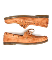 The Bed Stu Uncle Floyd tan leather boat shoes feature brown soles, lace detailing, and visible stitching in a classic nautical style, shown side-profile with one hand-finished shoe positioned above the other.