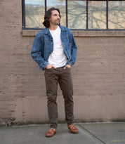 A man stands on a sidewalk in a denim jacket, white t-shirt, and brown pants, wearing Bed Stu’s Uncle Floyd hand-finished leather boat shoes with classic moc-toe stitching in front of a brick wall and large window.