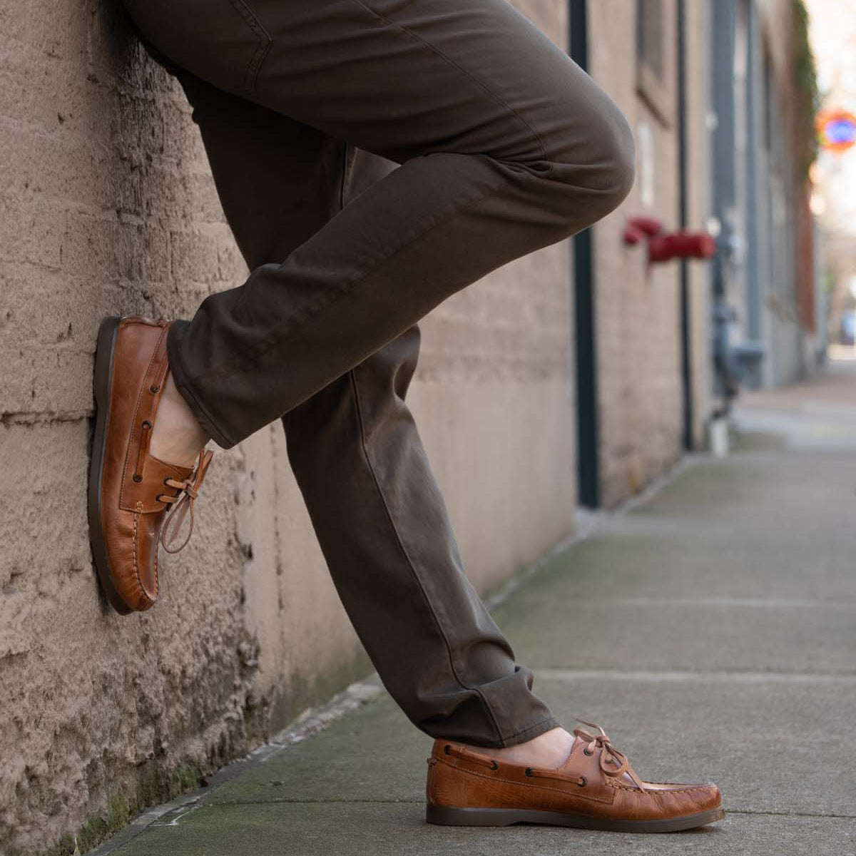 Wearing Bed Stu Uncle Floyd brown pants and tan leather boat shoes, a person leans against an outdoor wall on a city sidewalk.
