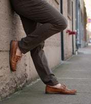 Wearing Bed Stu Uncle Floyd brown pants and tan leather boat shoes, a person leans against an outdoor wall on a city sidewalk.