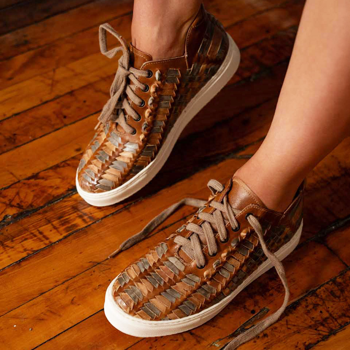 A person wearing Bed|Stü Unwavering handwoven leather sneakers with white soles stands on a wooden floor.
